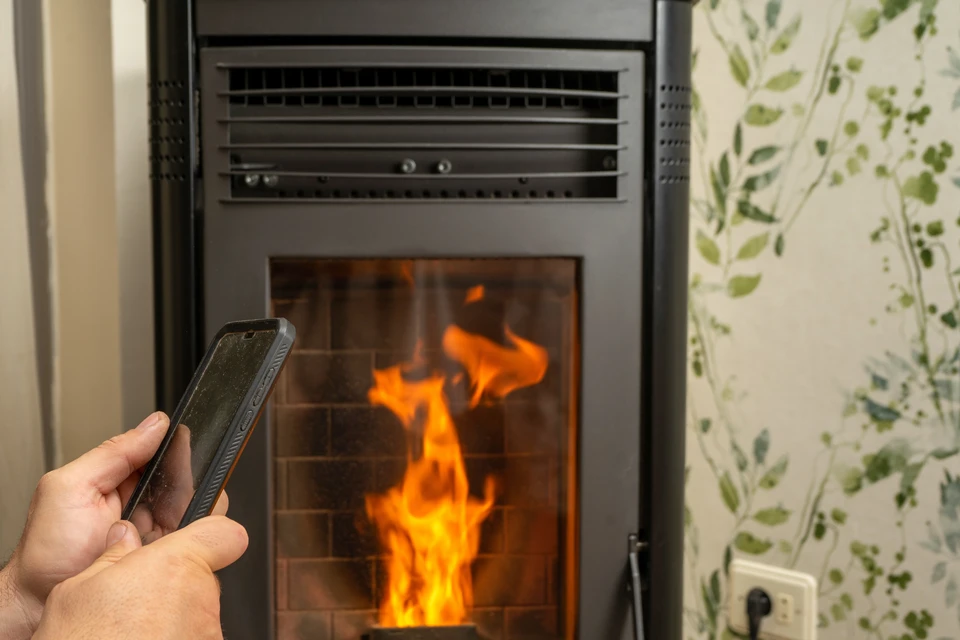 Technicien en tenue professionnelle effectuant l'installation d'un poêle à granulés design dans un salon, en train de raccorder le conduit d'évacuation métallique à l'arrière de l'appareil. Le poêle cylindrique noir est allumé, flammes visibles, installé dans un intérieur moderne et épuré. Ambiance de confiance et professionnalisme.